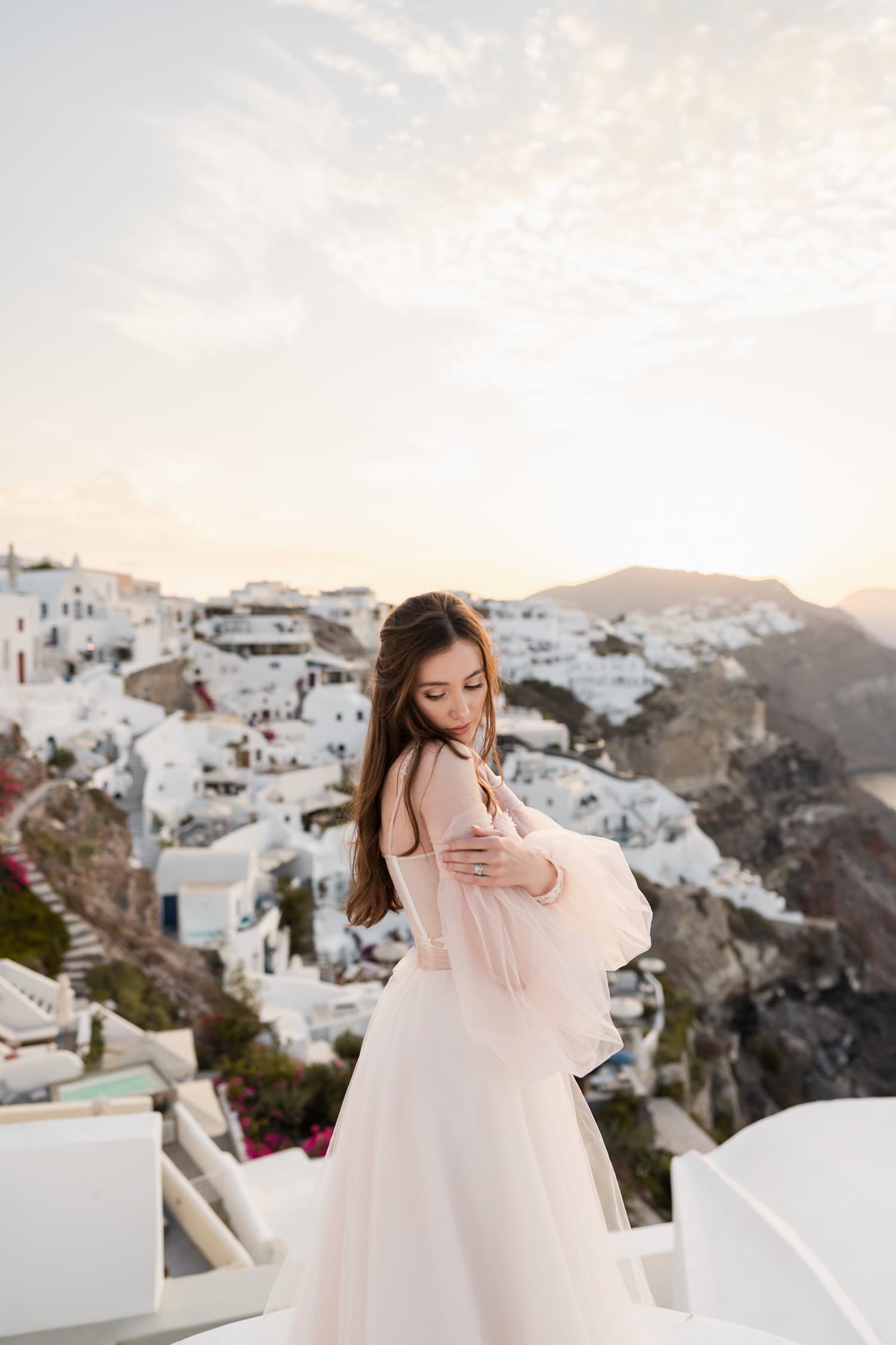 Bride possing on the top of a dome in Oia, Santorini during sunset. She's crossing her hands and has white houses in the background.