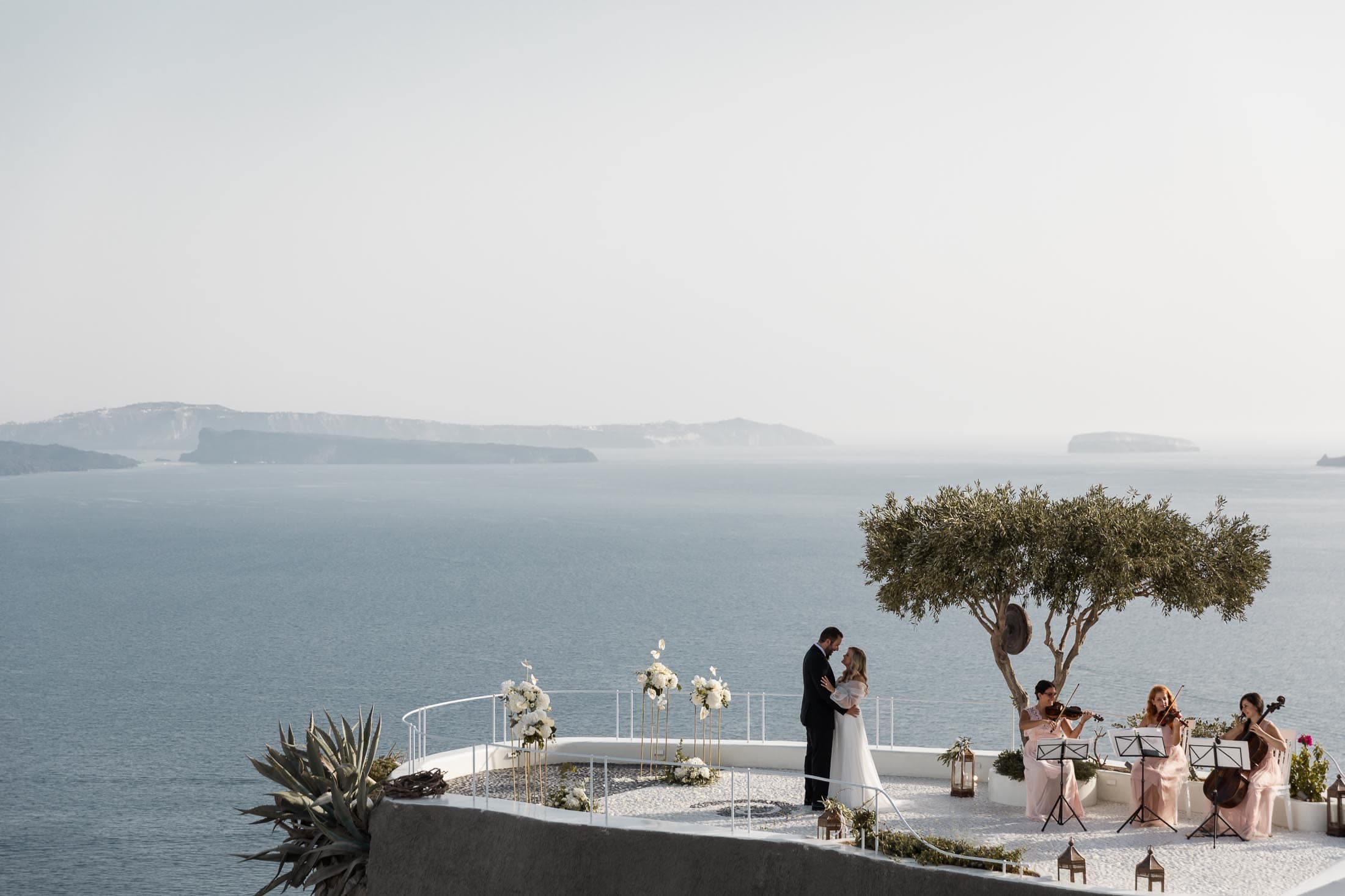 Wedding ceremony at Andronis luxury suites in Santorini. A string trio plaing music while the couple kisses.