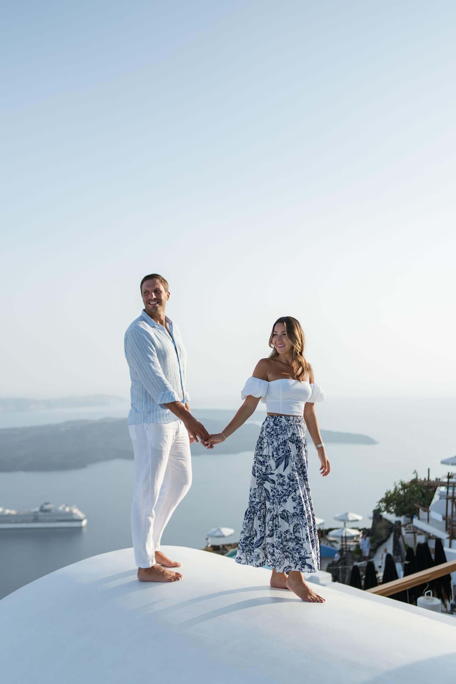 Coupla walking barefoot on a white dome in Santorini. They are posing for their private photo session.