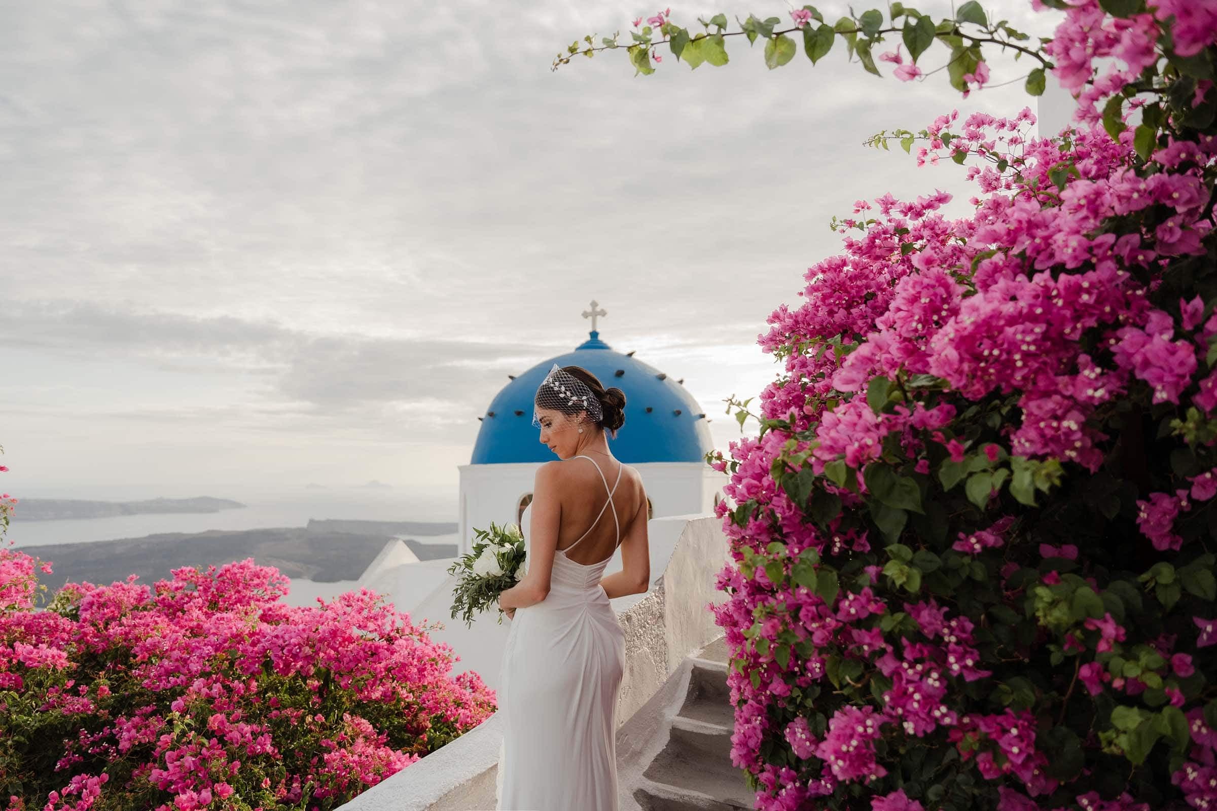 Elegant bride posing against the blue dome of Anastasi Church in Santorini. She is surrounded by blossomed bougainvillea flowers.