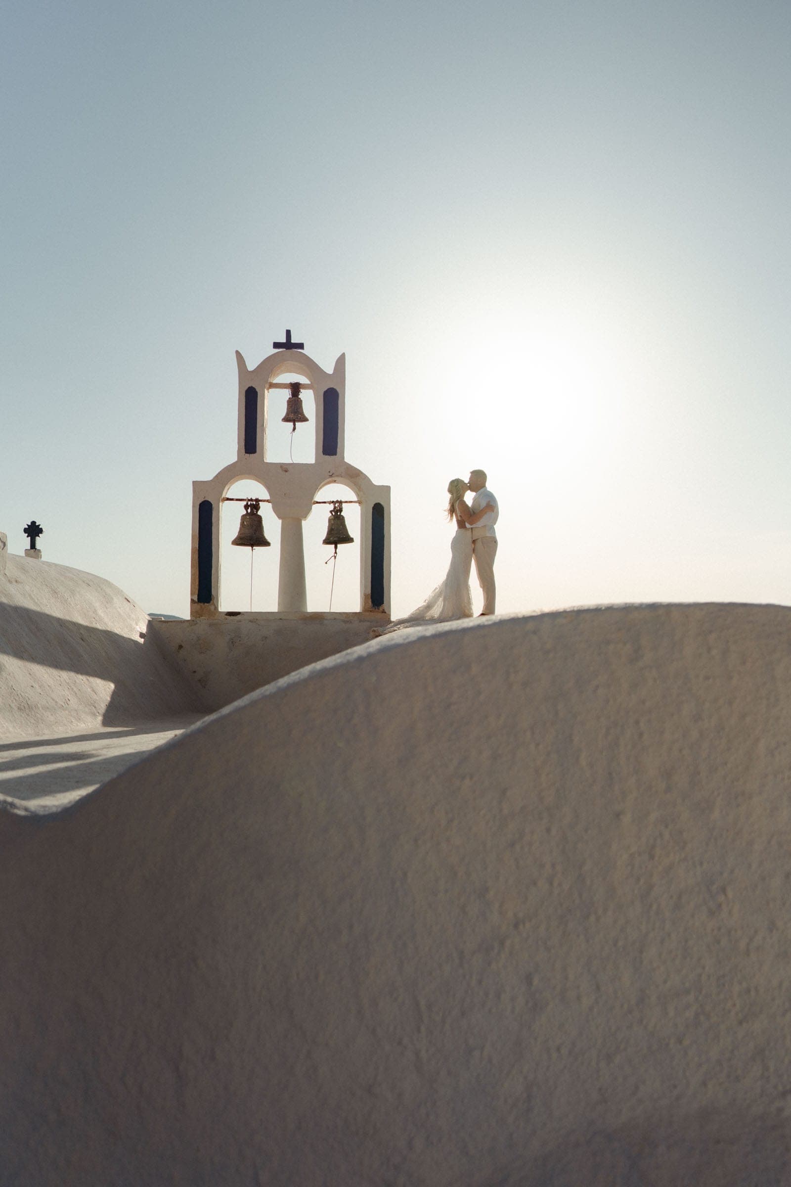 Wedding couple kissing on top of a three bell chapel in Santorini, Greece.