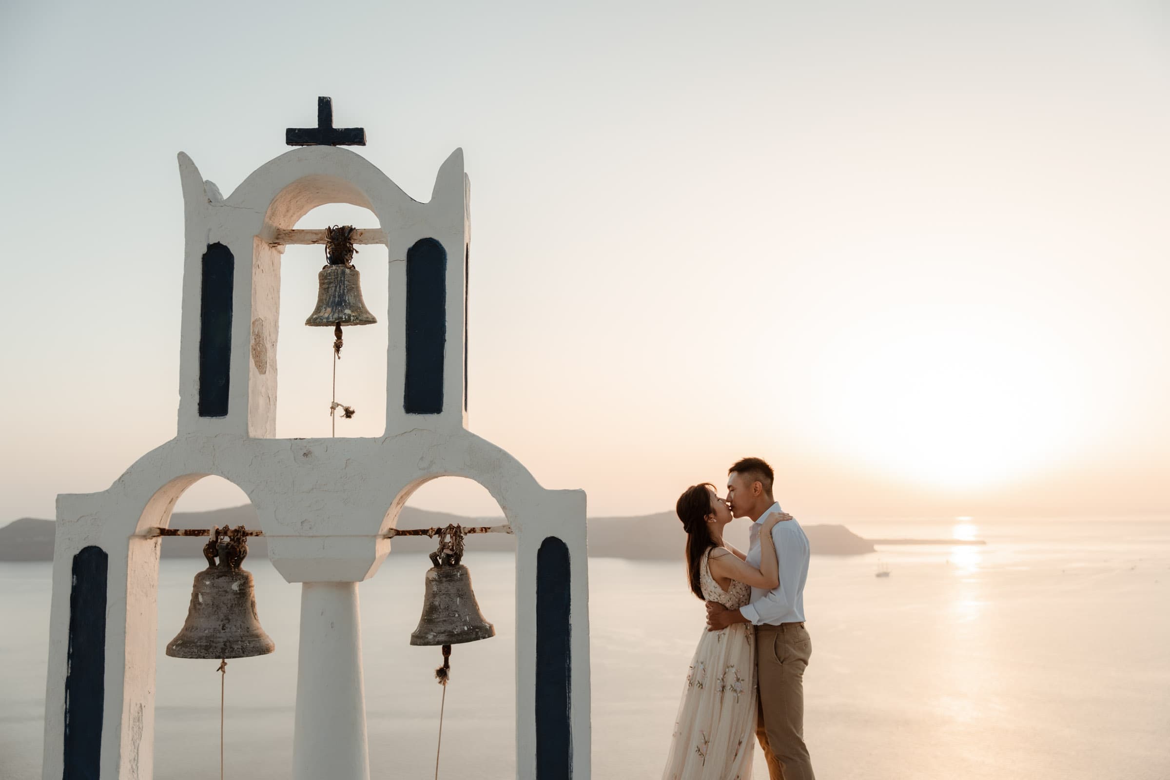 Couple kissing next to the three bell chapel in Santorini during sunset.