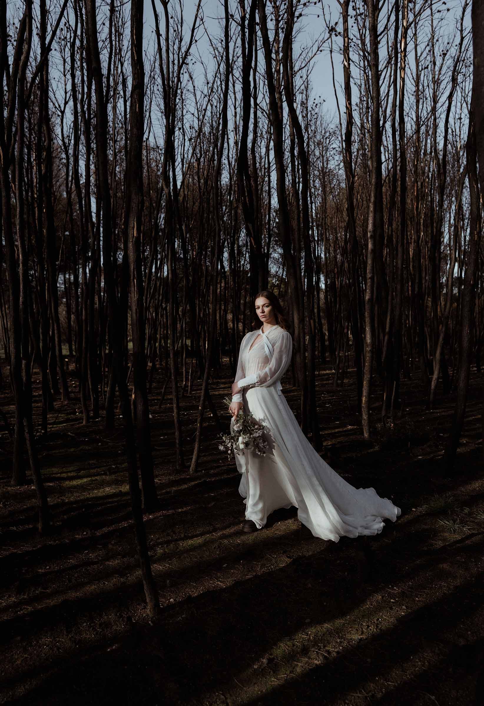 A bride in the forest holding her bridal bouquet. She is standing between burned out trees.